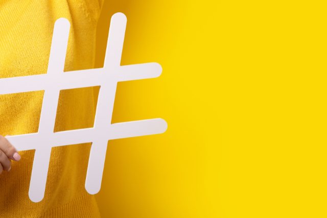 Person in a yellow top against a yellow background holding up a cutout of a hashtag